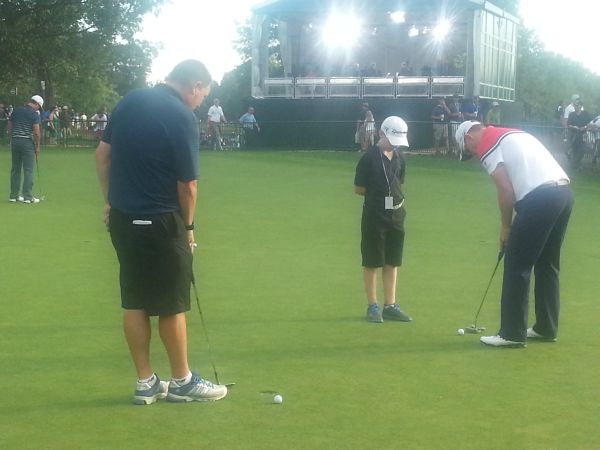 Young 12-year old Jack Gallacher stands watch over his father's putting like a coach as the lights from an on-course TV studio glow brightly. (Photo - www.golfbytourmiss.com)