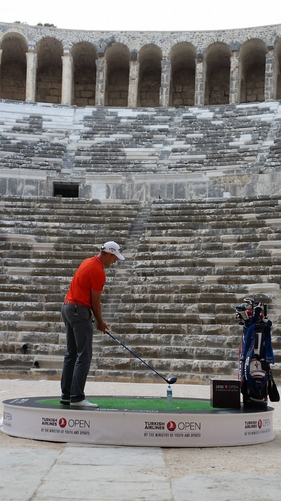 Henrik Stenson using driver to hit a golf ball off the top of a drink bottle within the Theatre of Aspendos